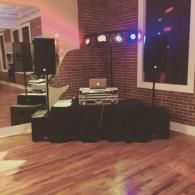 Small DJ setup with laptop and speakers on a black table in a brick-walled room with colored lights.