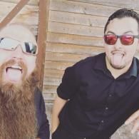 Two men wearing sunglasses laugh in front of a wooden wall.
