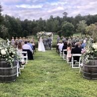 Outdoor wedding ceremony on a grassy aisle, with guests seated beside floral arrangements and wooden barrels.