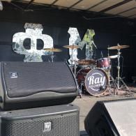 Empty concert stage with drum kit, speaker cabinets, and a backdrop logo reading “Ray.”
