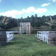 Outdoor wedding aisle with white chairs, wooden barrels, and a floral arch on a grassy lawn with trees in the background