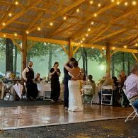Couple slow dancing on a wooden pavilion dance floor at a reception with string lights and seated guests