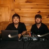 Two people seated at a table with audio equipment and a laptop against a wooden wall