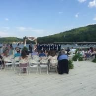 Outdoor wedding ceremony on a lakeside patio with rows of chairs, guests, and a floral arch against green hills.