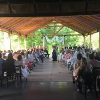 Guests seated at a decorated outdoor pavilion wedding ceremony with aisle and greenery backdrop