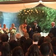 Wedding couple kissing under floral arch as guests and officiant look on indoors