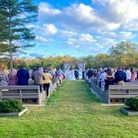 Crowd gathered on a grassy path toward a white arch outdoors under a blue sky with clouds.