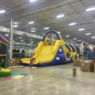 Large yellow-and-blue inflatable slide in a warehouse with high ceilings and stacked boxes