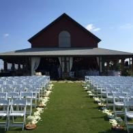 Outdoor wedding setup with rows of white chairs facing a barn-like venue under a blue sky