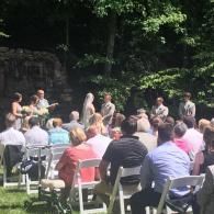 Outdoor ceremony with seated guests watching a speaker in a wooded setting