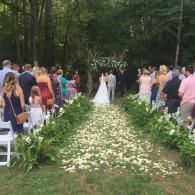 Outdoor wedding ceremony aisle lined with white flowers, guests seated on both sides in a forest clearing