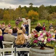 Outdoor wedding ceremony with floral arch, guests seated, and a large pink flower arrangement in the foreground