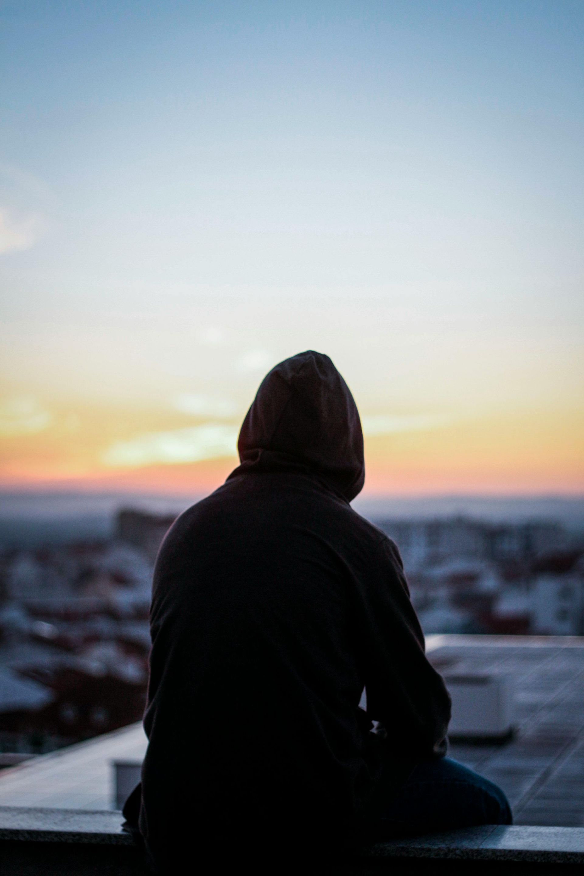 A man in a hoodie is sitting on a ledge looking at the sunset.