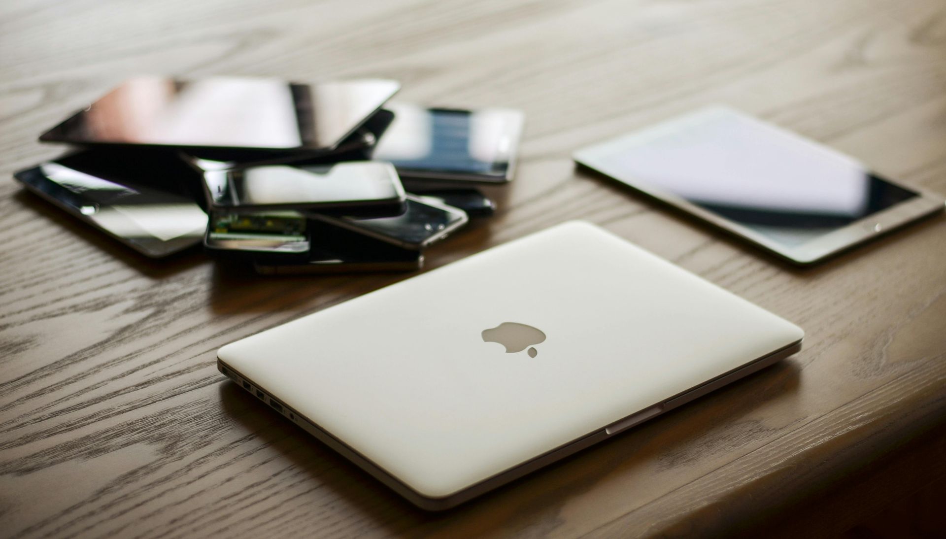 An apple laptop is sitting on a wooden table next to a stack of cell phones and a tablet.
