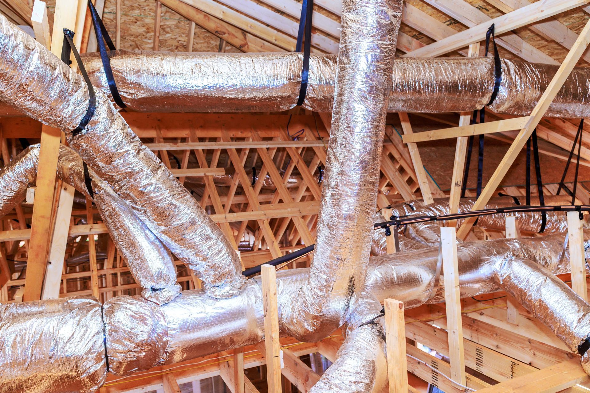 Exposed attic framing with multiple silver HVAC ducts and vents running through wooden roof trusses