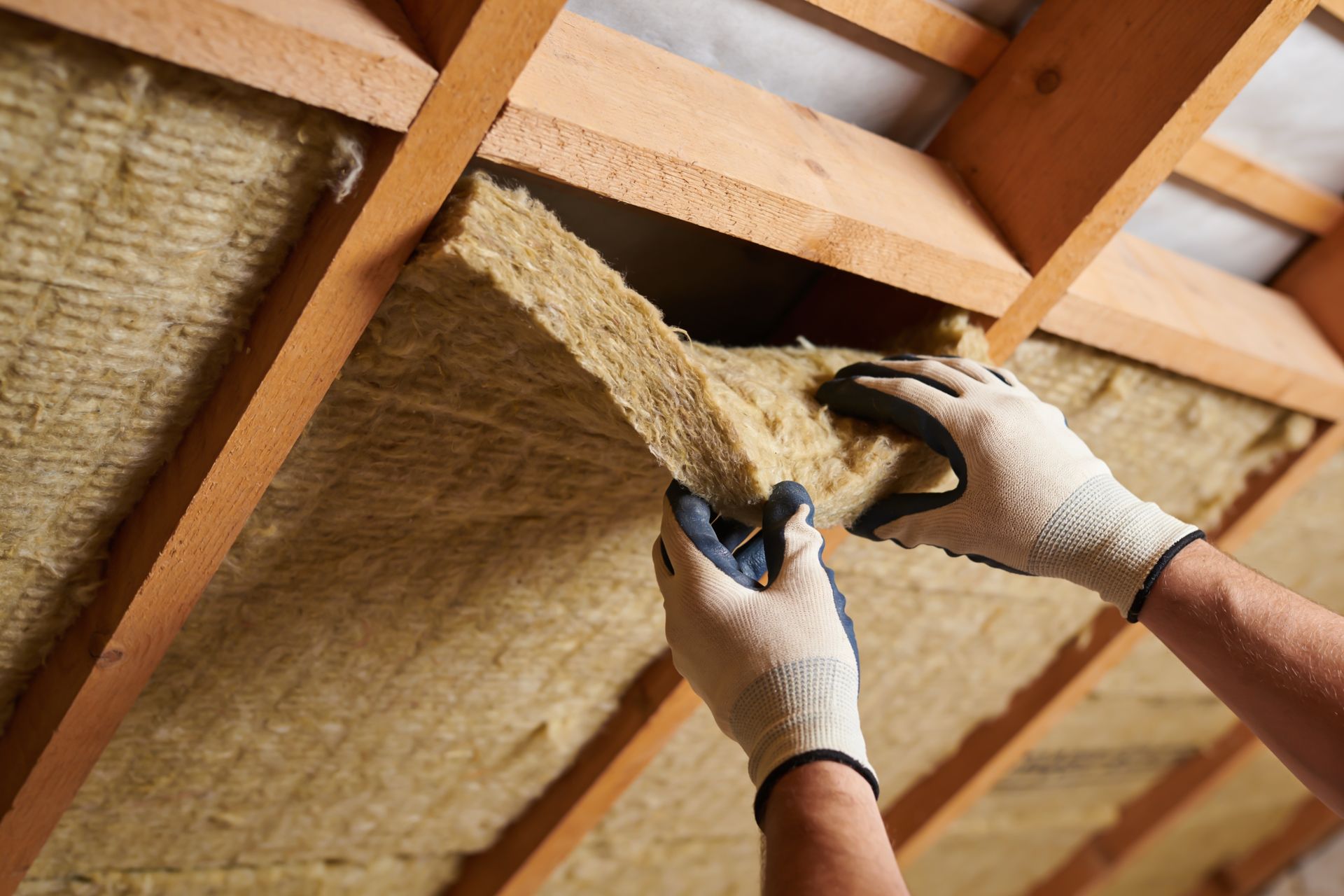 Gloved hands installing insulation batts between wooden roof rafters