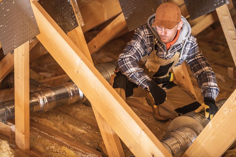Worker inspecting attic framing with exposed wooden beams and insulation, wearing plaid shirt and hard hat