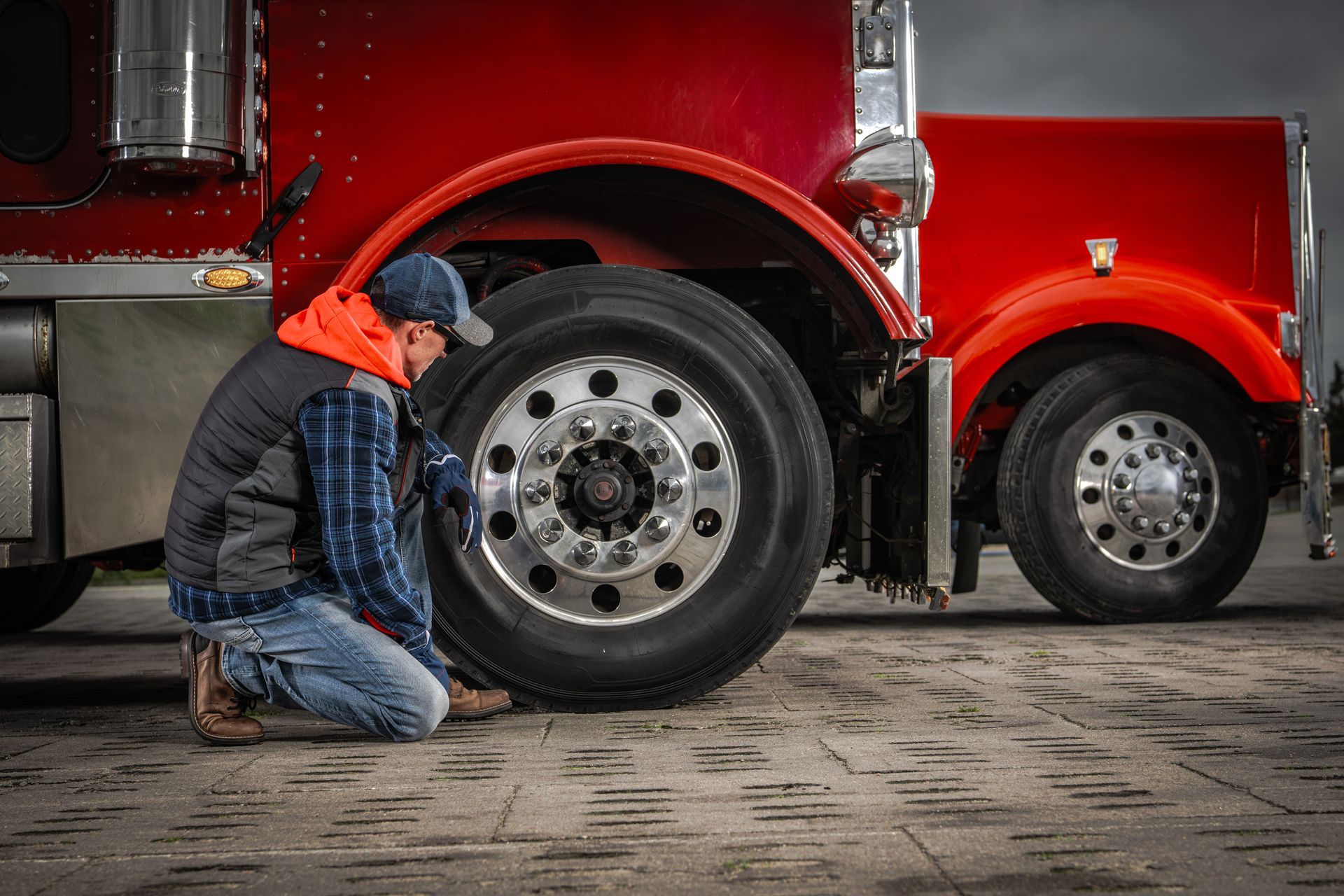 Person kneels to check a semi-truck tire; red truck, brick pavement, overcast sky. Person kneels to check a semi-truck tire; red truck, brick pavement, overcast sky.