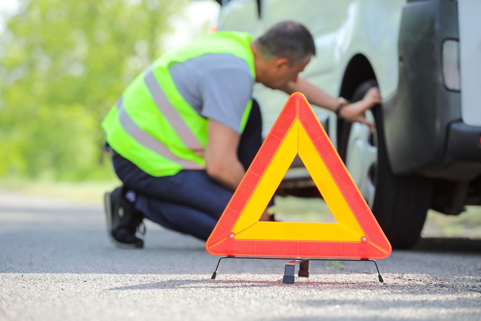 Man in safety vest checks tire, warning triangle on road. Man in safety vest checks tire, warning triangle on road.
