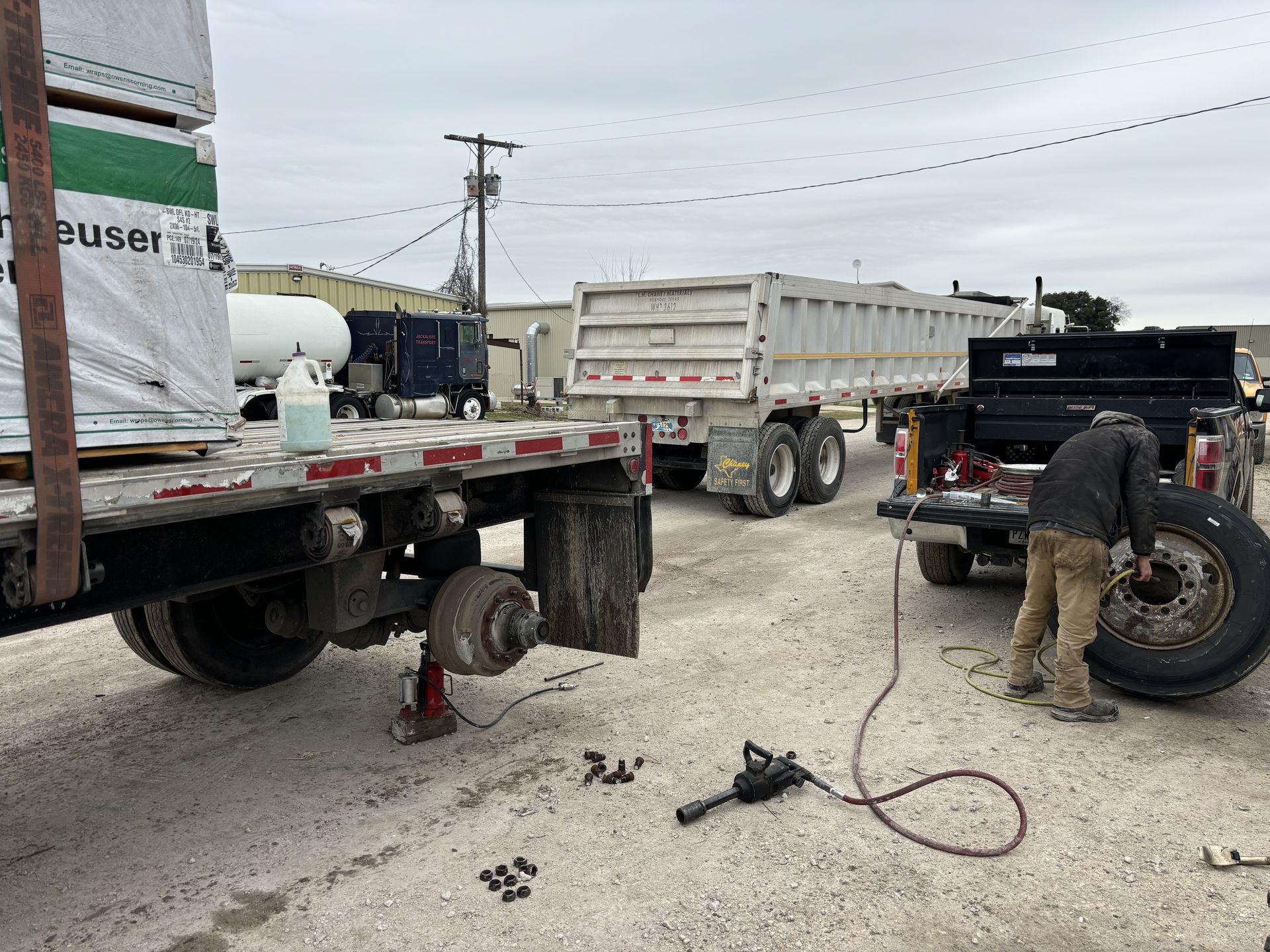 A person changes a tire on a flatbed trailer, another trailer and truck are nearby, on a cloudy day. A person changes a tire on a flatbed trailer, another trailer and truck are nearby, on a cloudy day.
