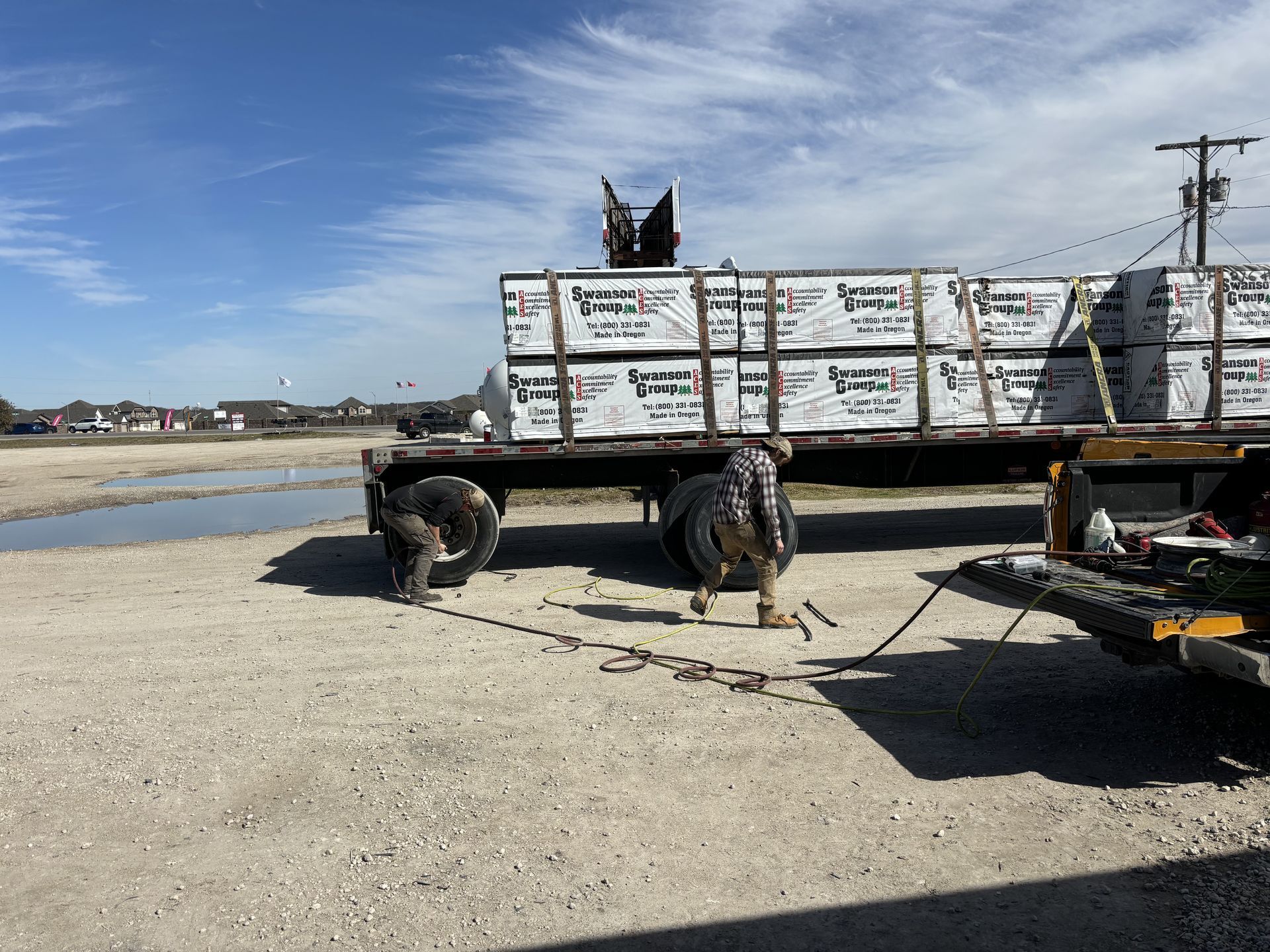 Flatbed trailer loaded with lumber being secured by workers in a gravel lot on a sunny day. Flatbed trailer loaded with lumber being secured by workers in a gravel lot on a sunny day.