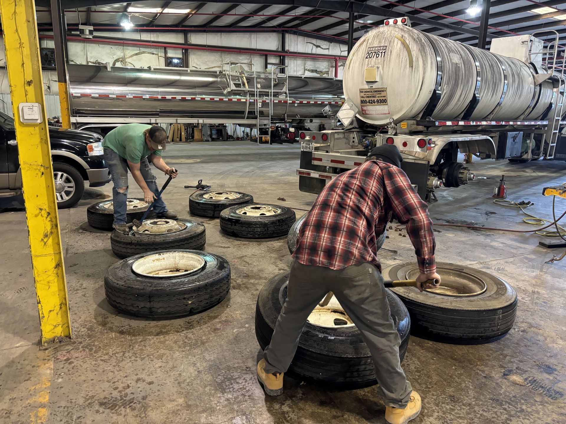 Two workers changing truck tires in a shop with a tanker truck in the background. Two workers changing truck tires in a shop with a tanker truck in the background.