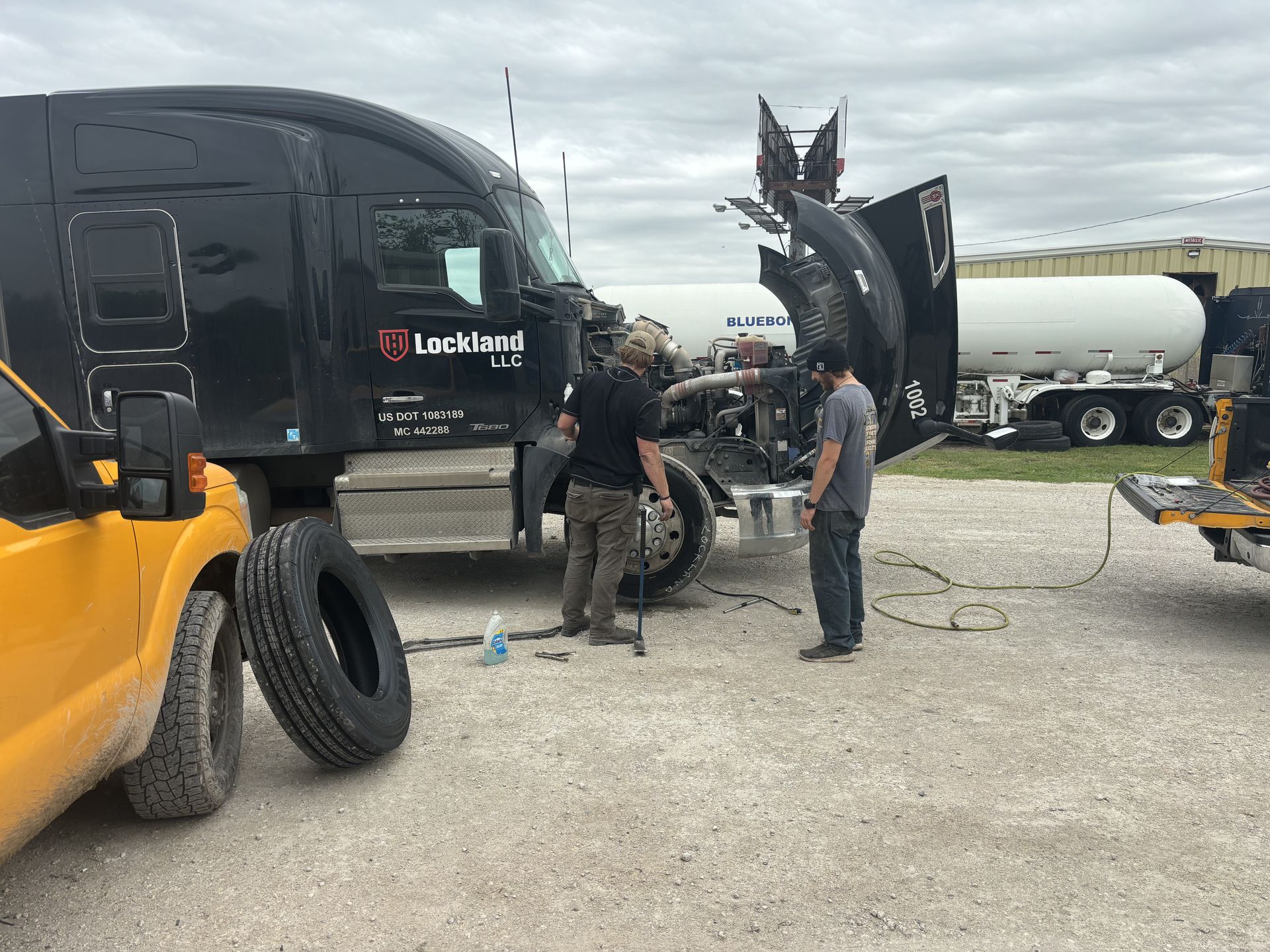 Two people working on a black semi-truck with open hood. Yellow vehicle, spare tire, and propane tank visible in a gravel lot. Two people working on a black semi-truck with open hood. Yellow vehicle, spare tire, and propane tank visible in a gravel lot.