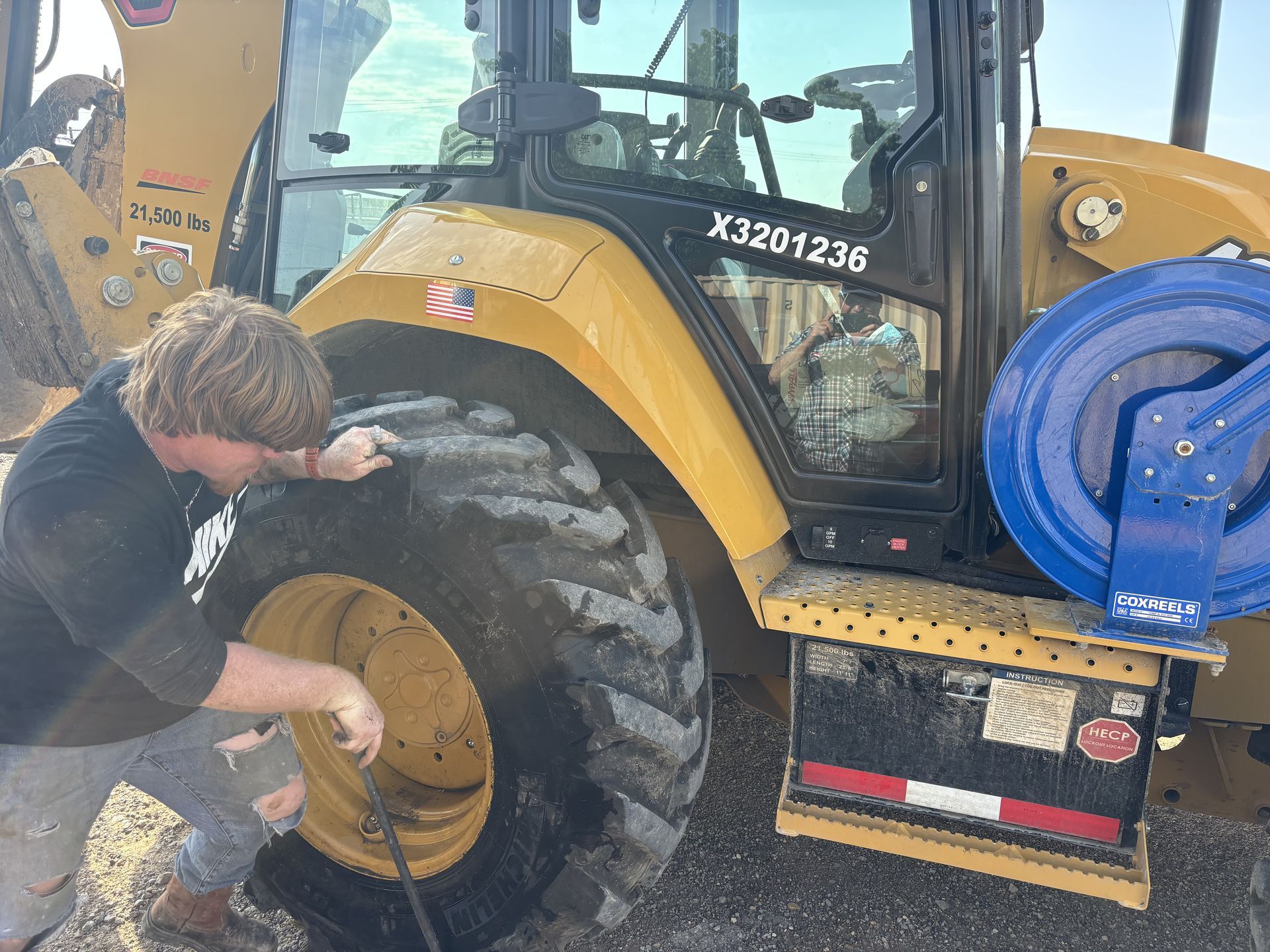 Man checks the tire of a yellow Caterpillar backhoe. The backhoe has a blue attached element and the man is wearing a black t-shirt. Man checks the tire of a yellow Caterpillar backhoe. The backhoe has a blue attached element and the man is wearing a black t-shirt.