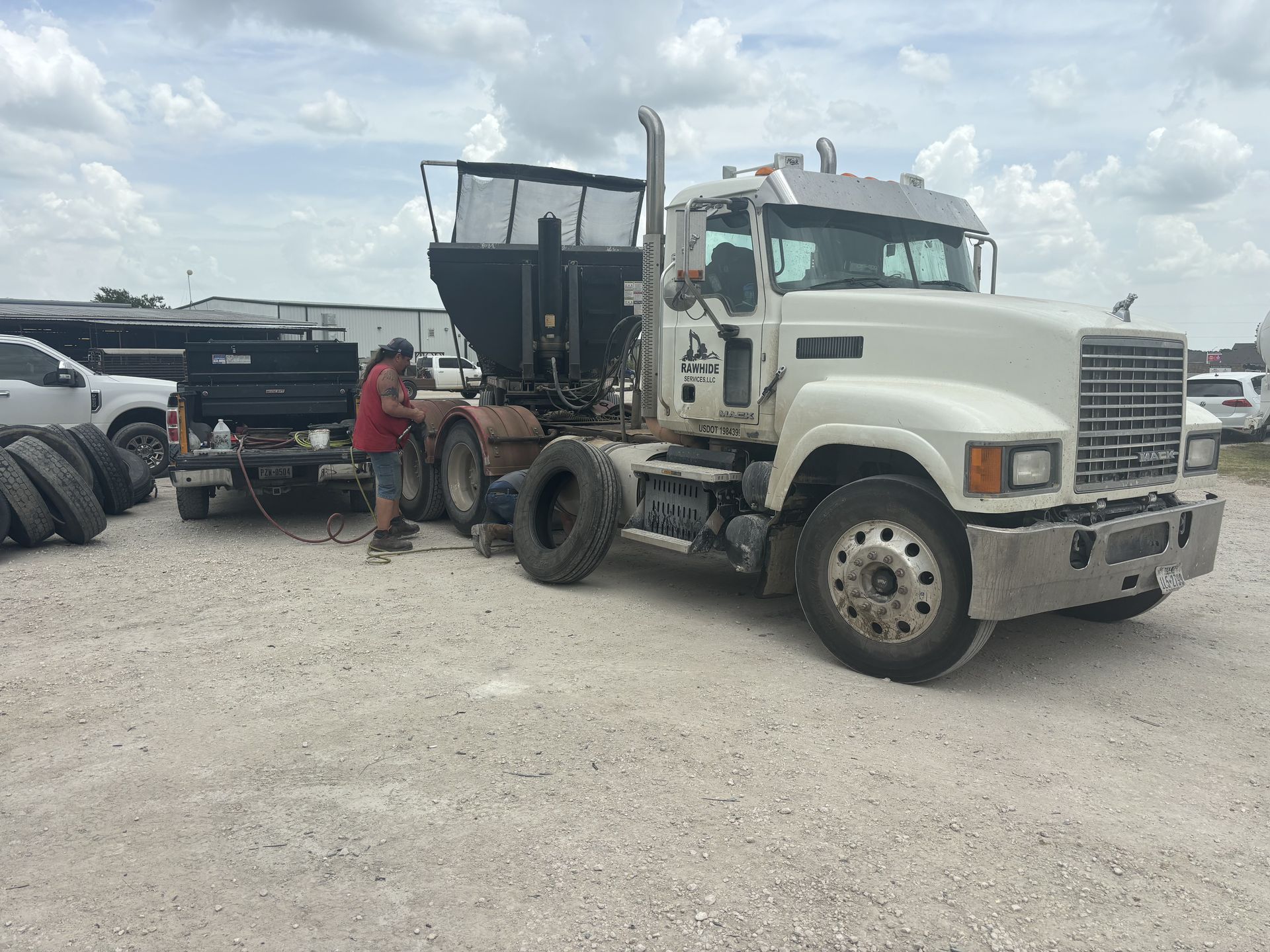 White Mack semi-truck with a black trailer; a person works on the truck in a gravel lot on a cloudy day. White Mack semi-truck with a black trailer; a person works on the truck in a gravel lot on a cloudy day.