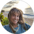 Smiling woman in a blue shirt and necklace standing outdoors by a coastal overlook.