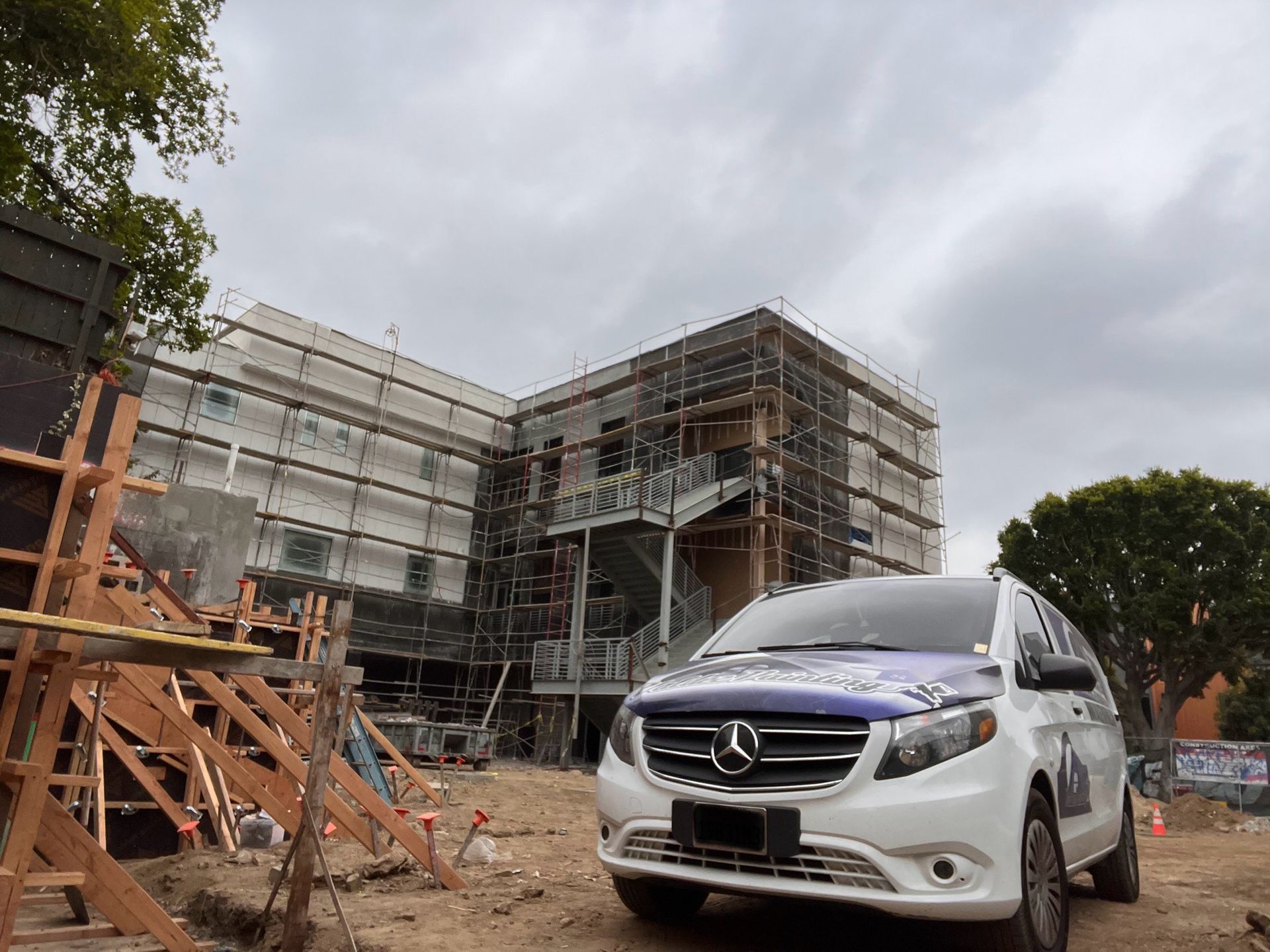 A white Mercedes-Benz van parked at a construction site with scaffolding around a building under a cloudy sky.