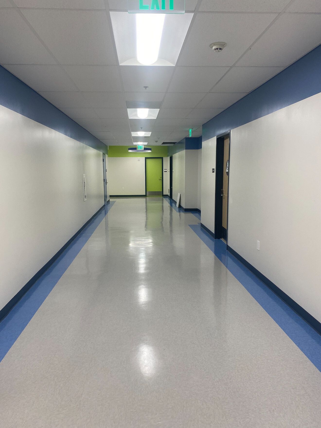 A long, empty indoor hallway with white walls, blue trim, a shiny grey floor, and green accent walls at the end.