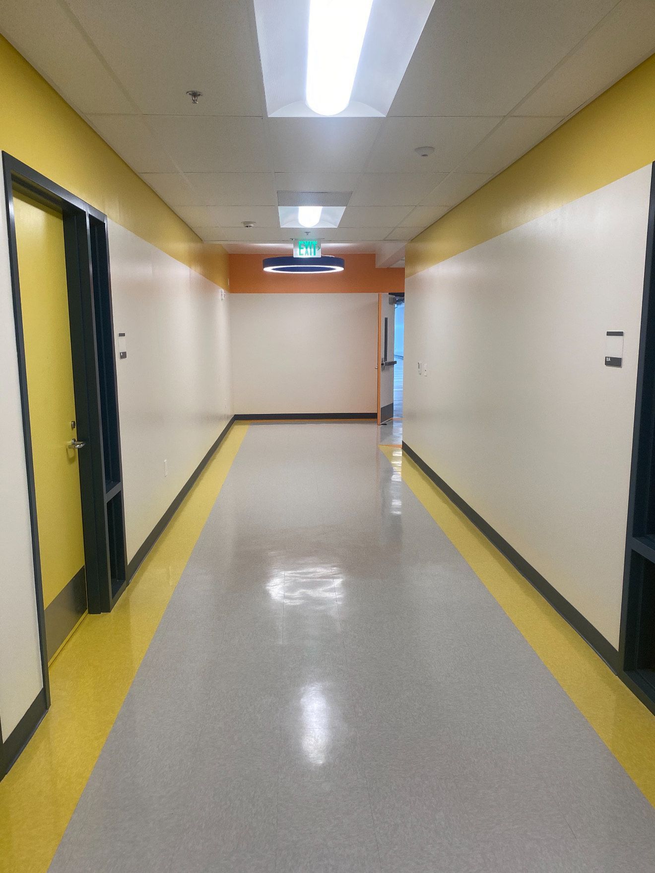 A straight, empty indoor hallway with white walls, yellow floor trim, and overhead lights leading to a green exit sign.