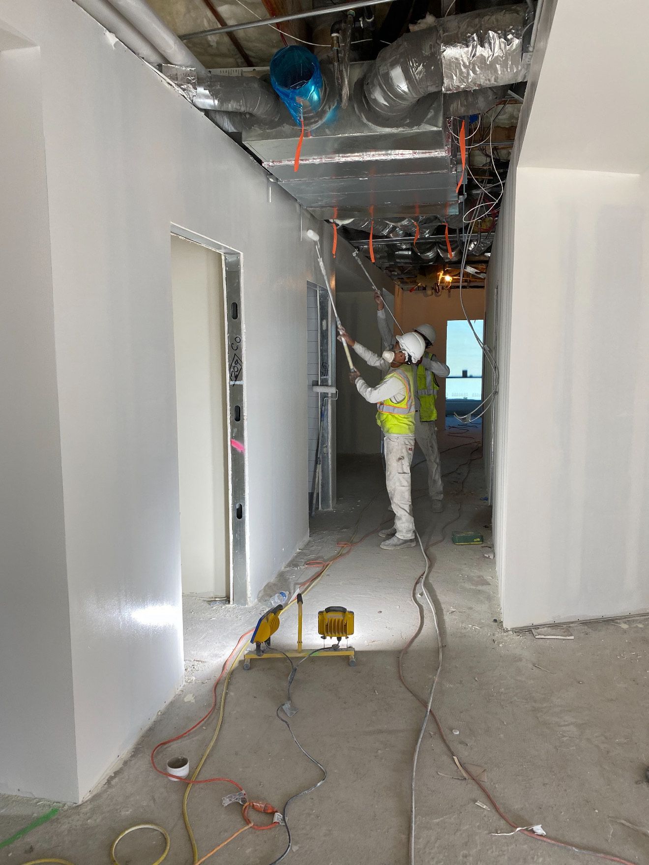 A worker in a high-visibility vest and hard hat uses a paint roller on the ceiling in an unfinished building corridor.