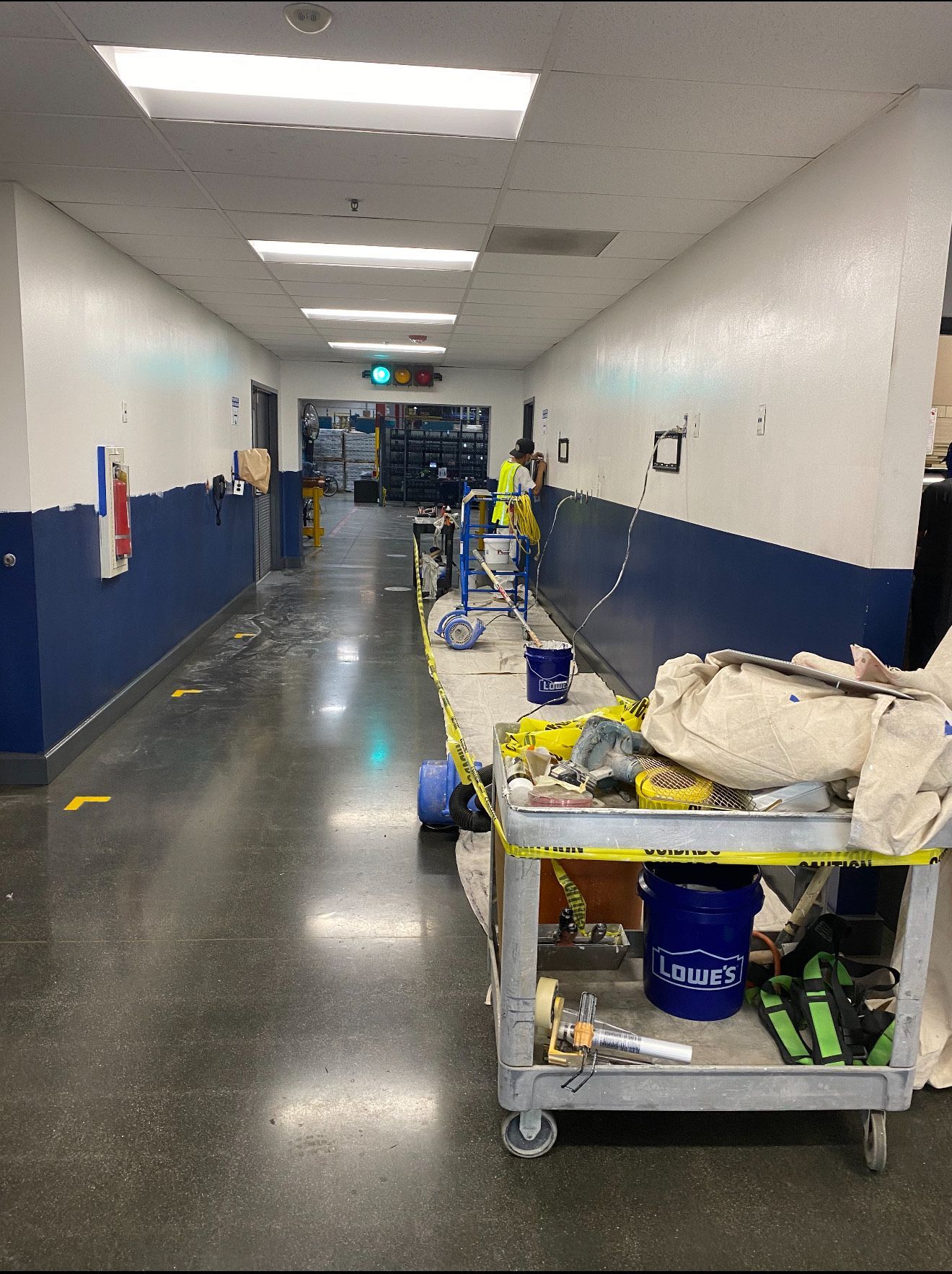 A worker paints a hallway with white and navy blue walls, featuring a utility cart and caution tape in the foreground.