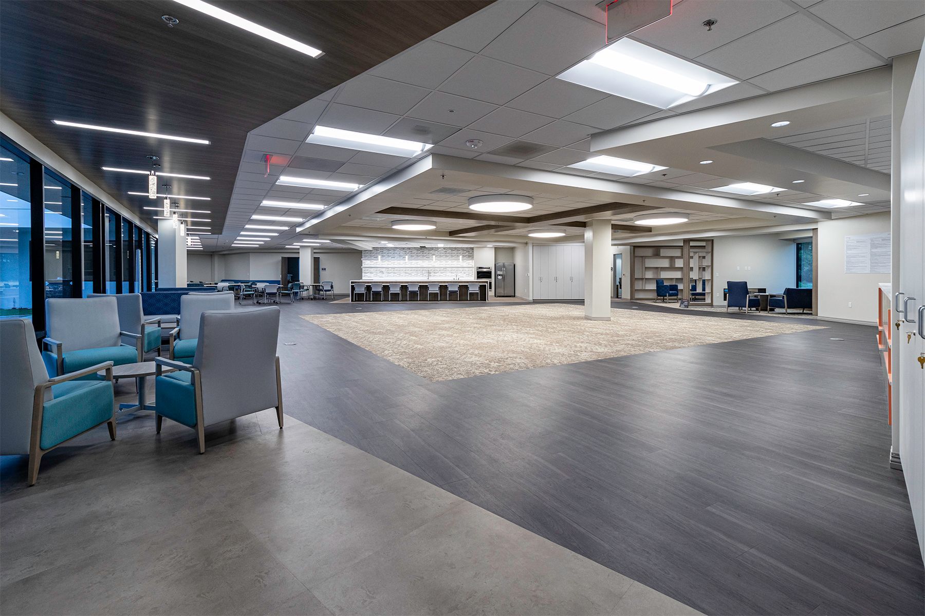 A spacious, modern office lounge with dark wood flooring, blue armchairs, recessed ceiling lights, and a white pillar.