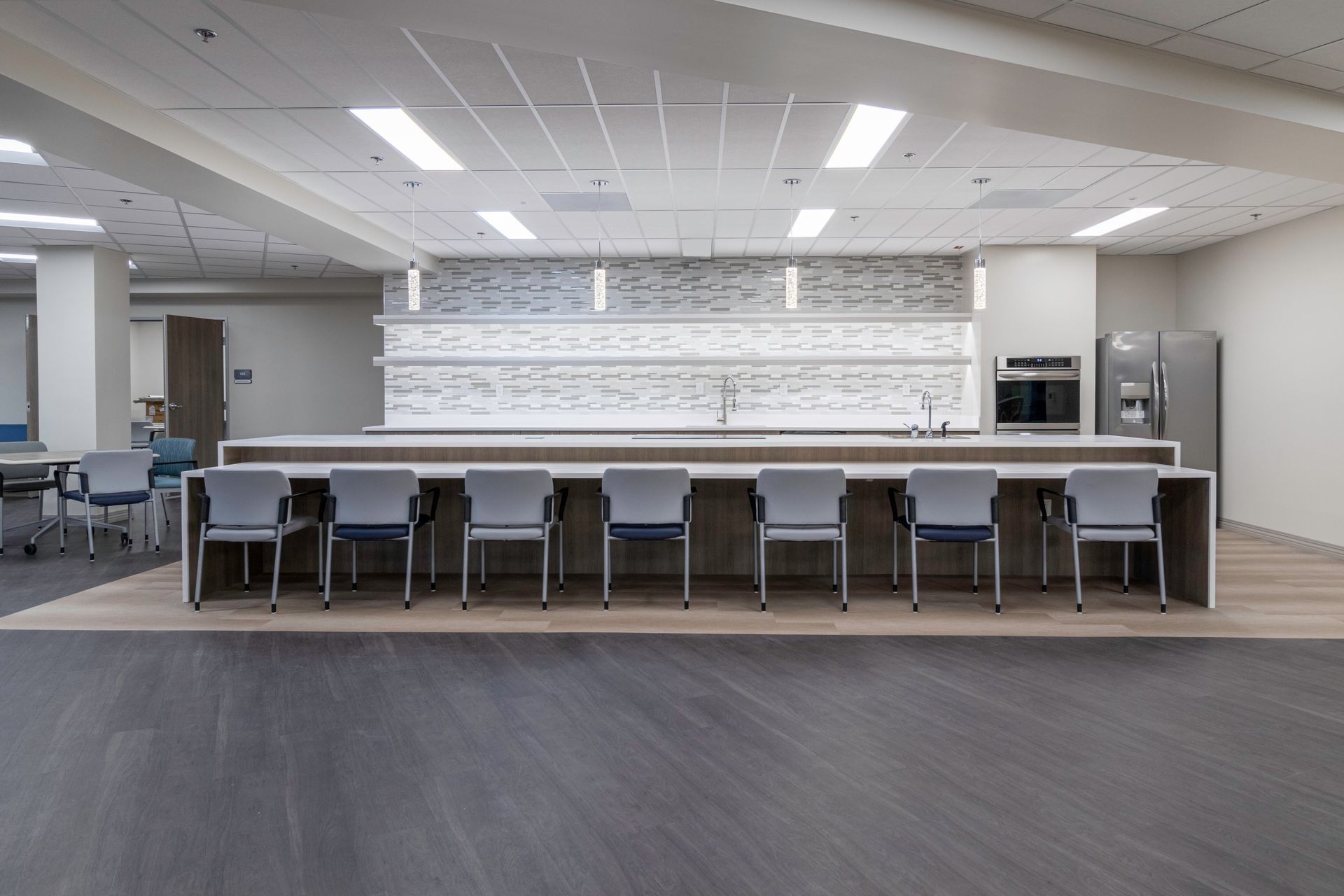 A modern office breakroom with a long, white counter, seven gray chairs, an oven, and a refrigerator against a tiled wall.