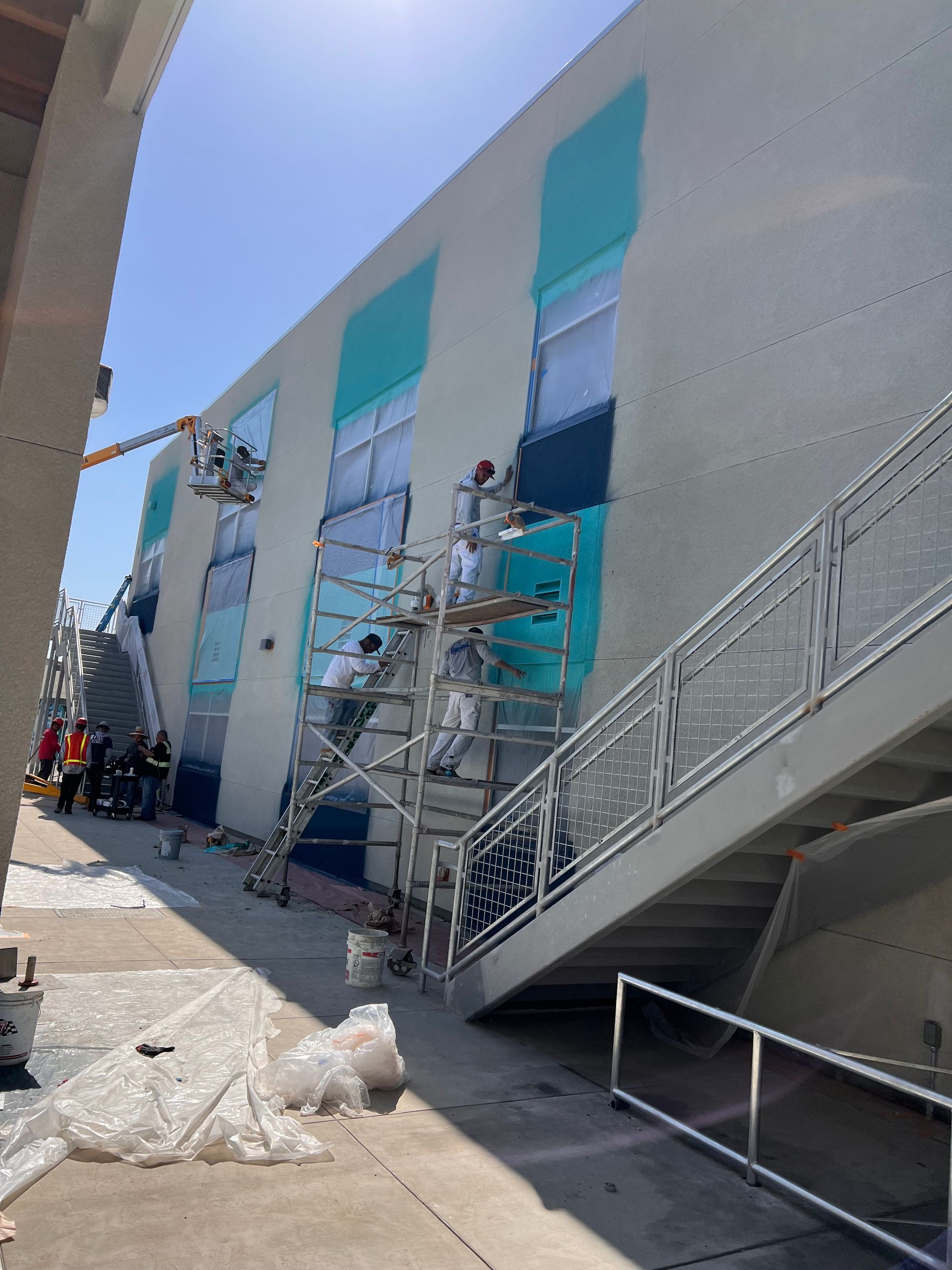 Workers use scaffolding and a lift to paint bright blue rectangles on the side of a tall, beige building.