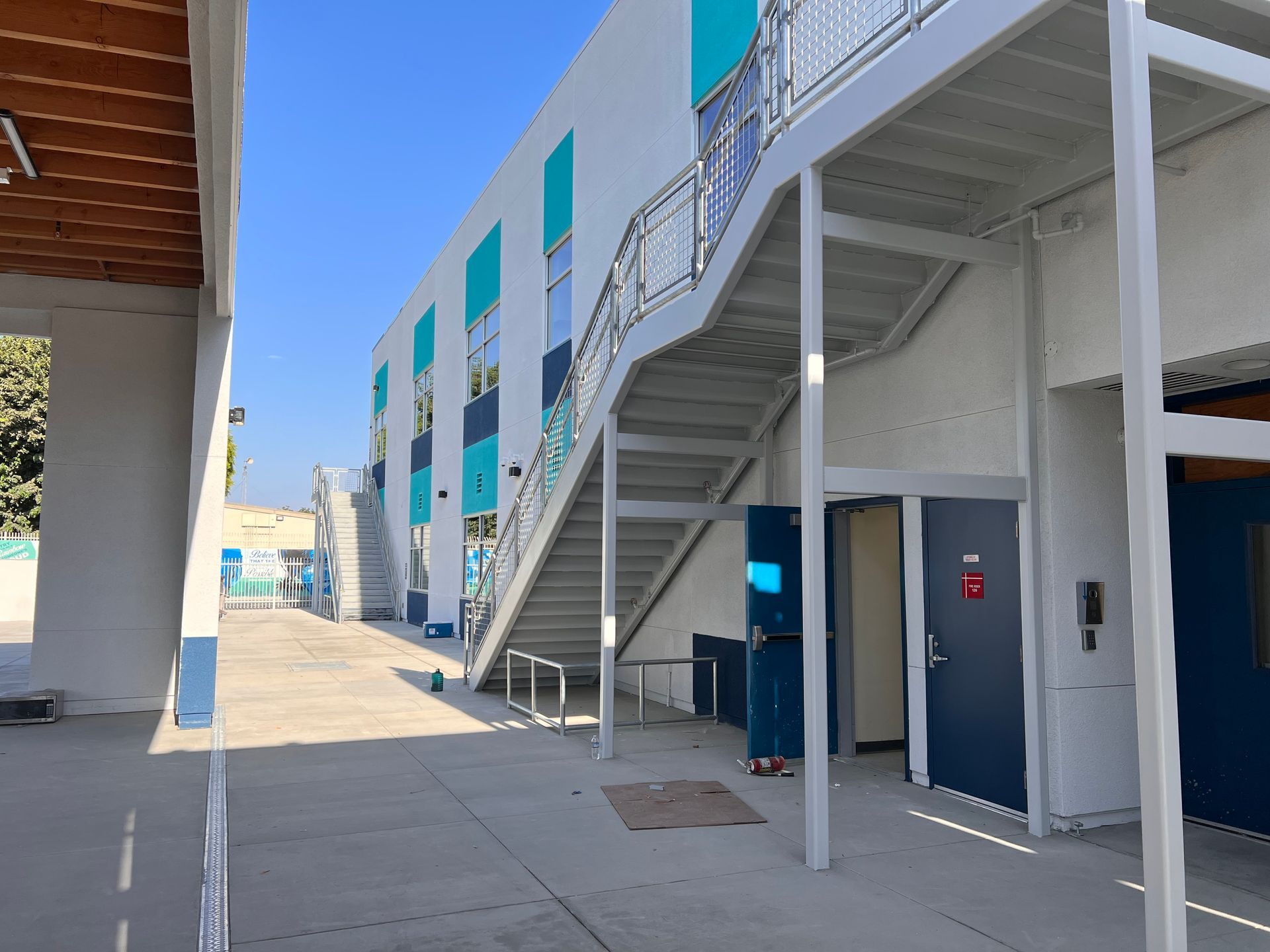 An outdoor walkway between two modern buildings with blue and white accents, featuring metal staircases and doorways.
