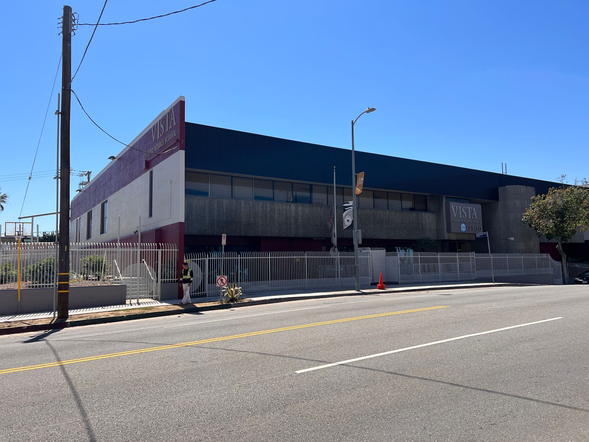 A two-story building with a white and maroon facade and a dark roof, behind a white metal fence along a street.