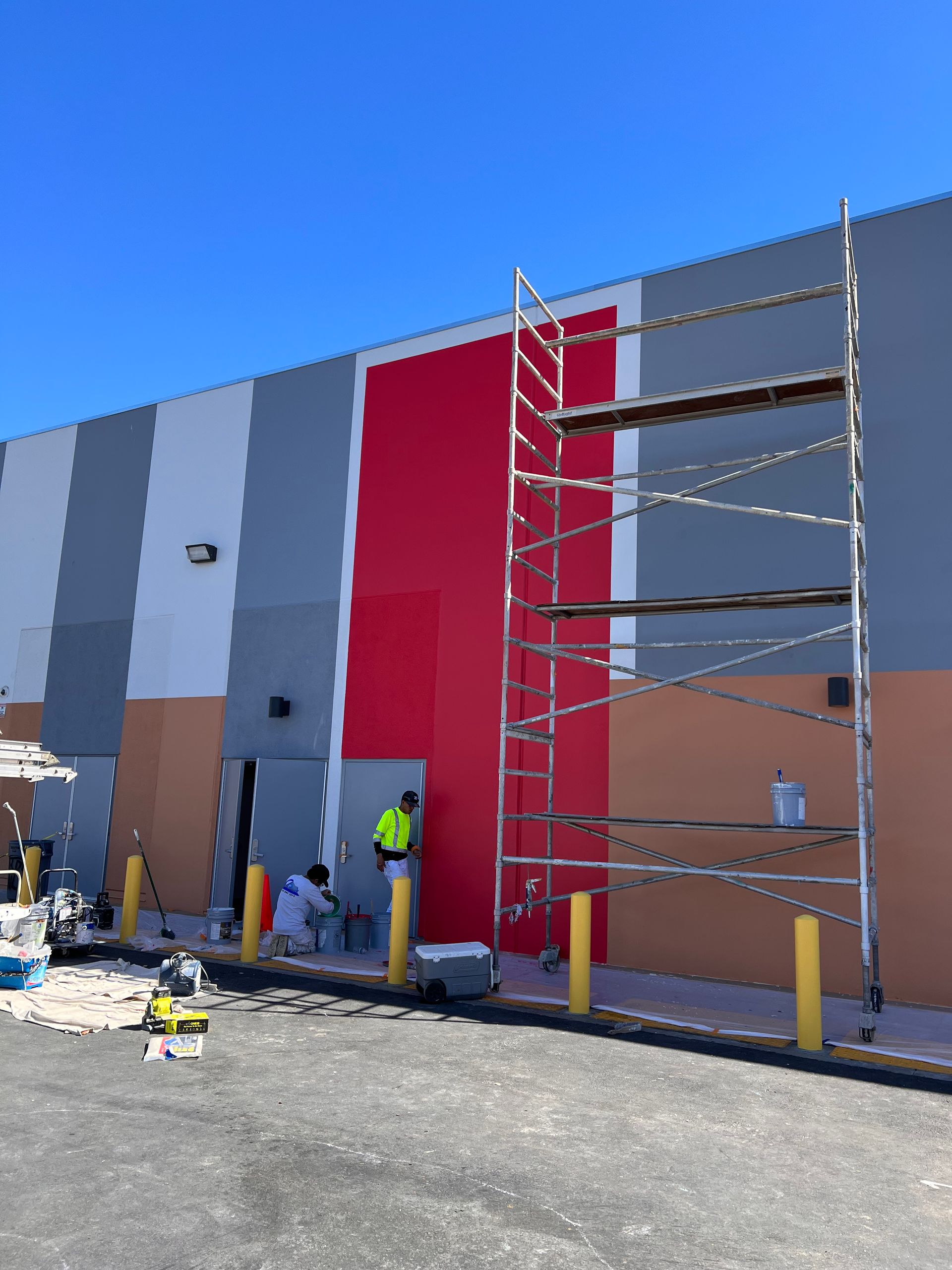Two workers paint a commercial building exterior with vertical stripes of white, gray, red, and tan under a blue sky.
