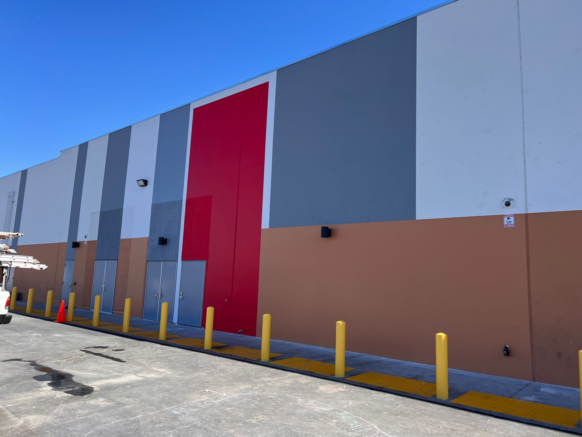Exterior of a warehouse building with gray, white, and red vertical panels above a brown base, lined with yellow bollards.