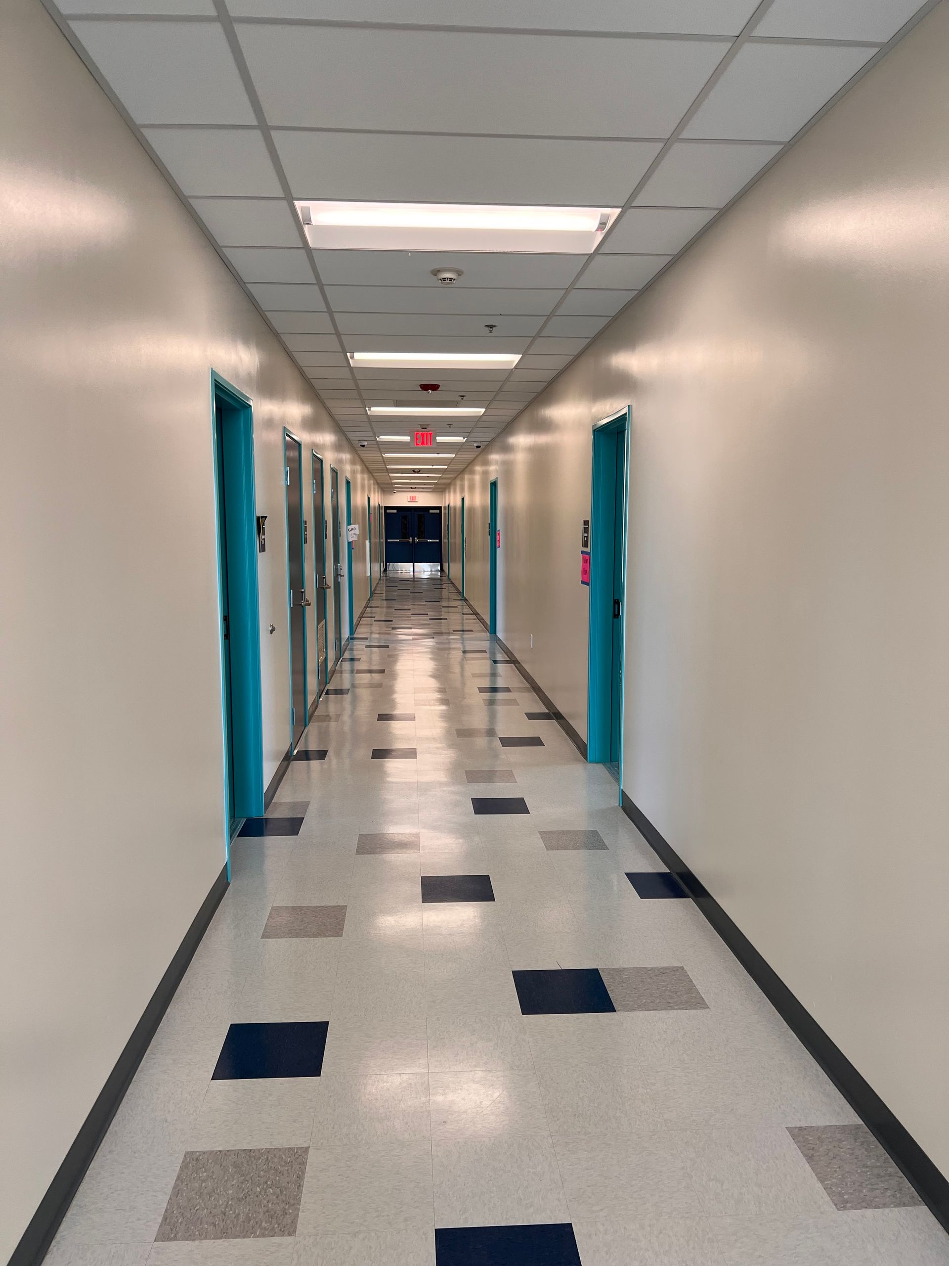 Long, symmetrical hallway with bright lights, light-colored walls, teal door frames, and a speckled floor with blue tiles.