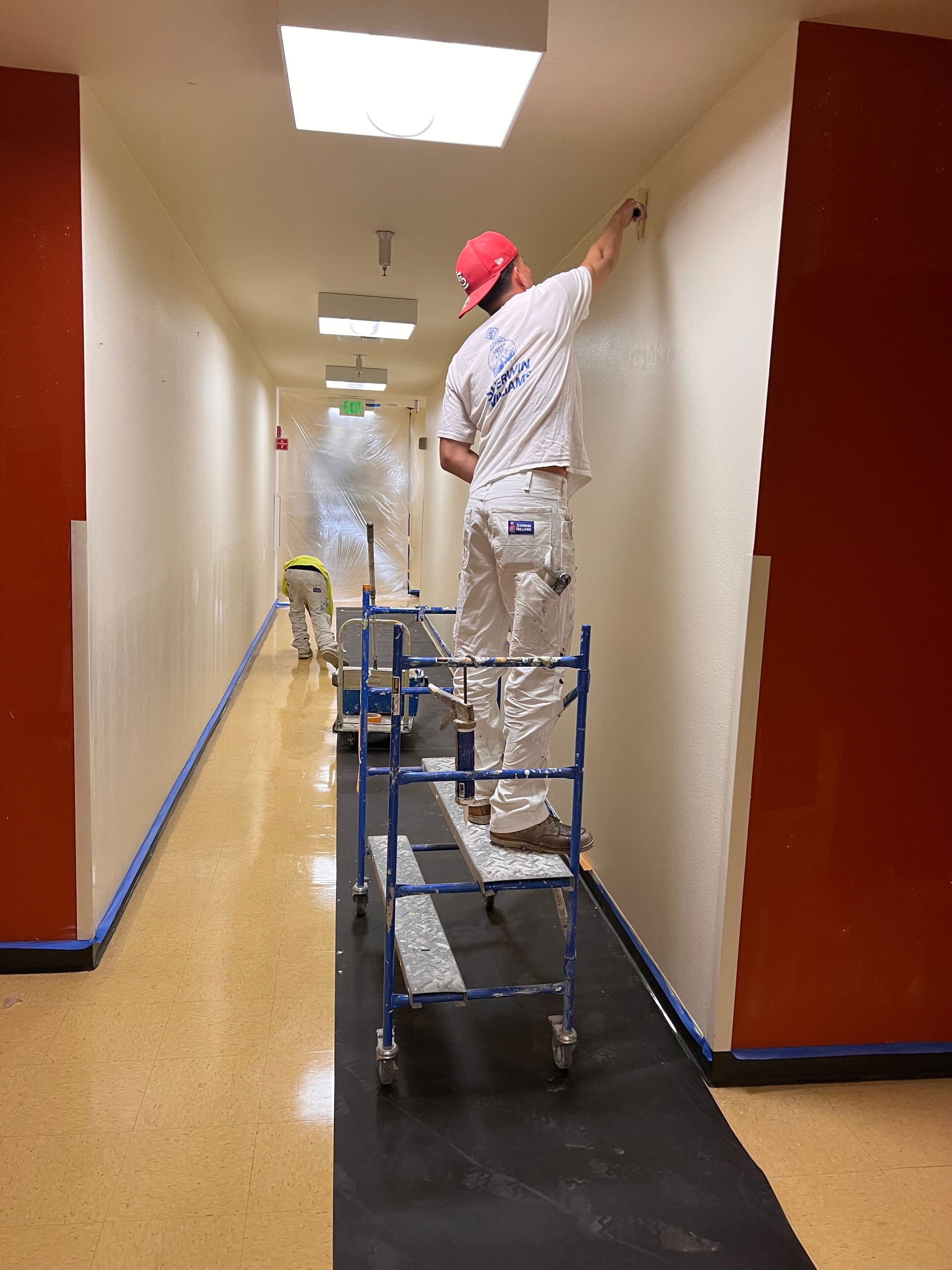 A painter in white clothing stands on a blue metal scaffold, painting an off-white hallway wall.