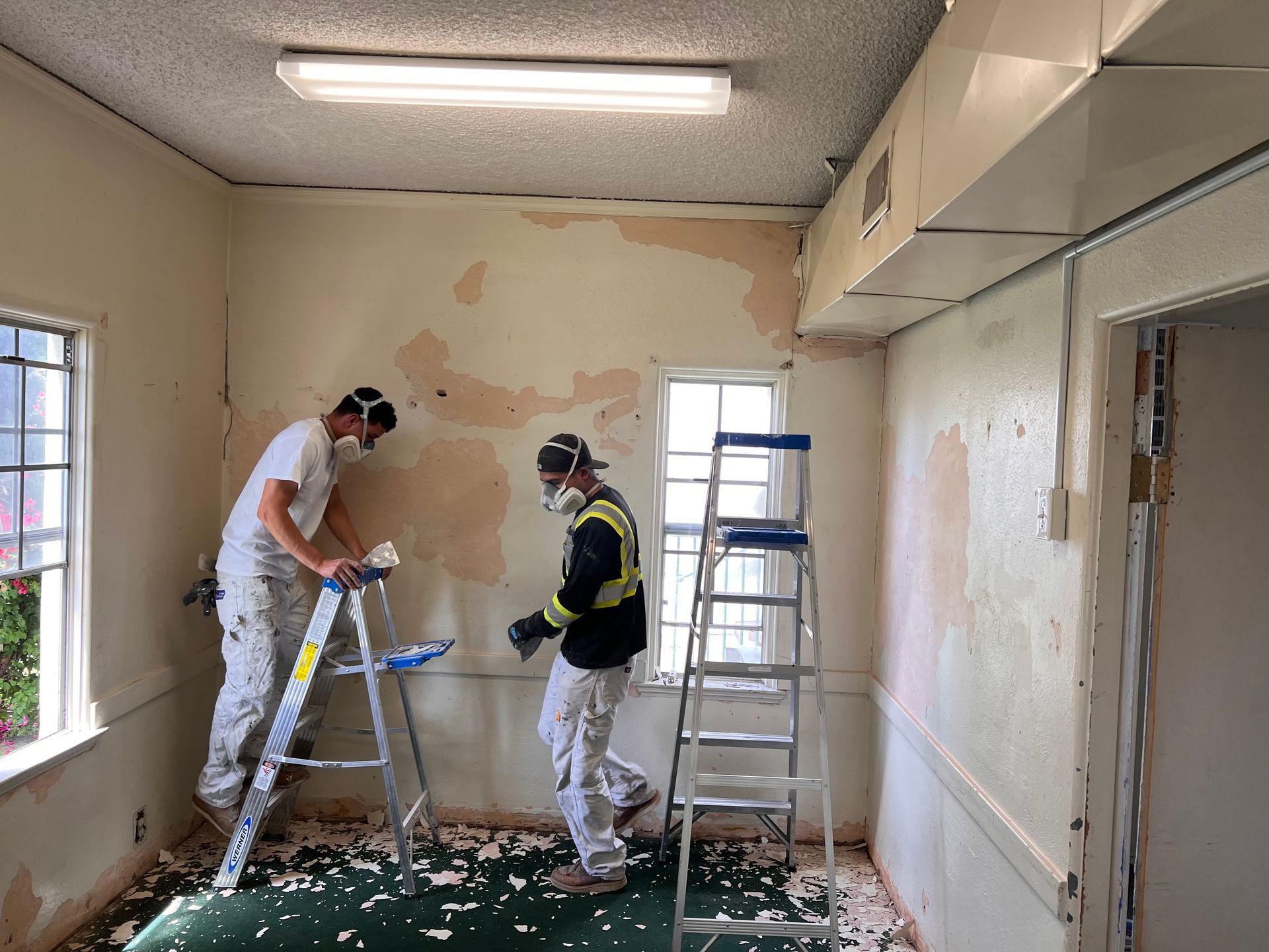Two people in protective gear and masks strip wallpaper in a room with a step ladder and debris-covered floor.