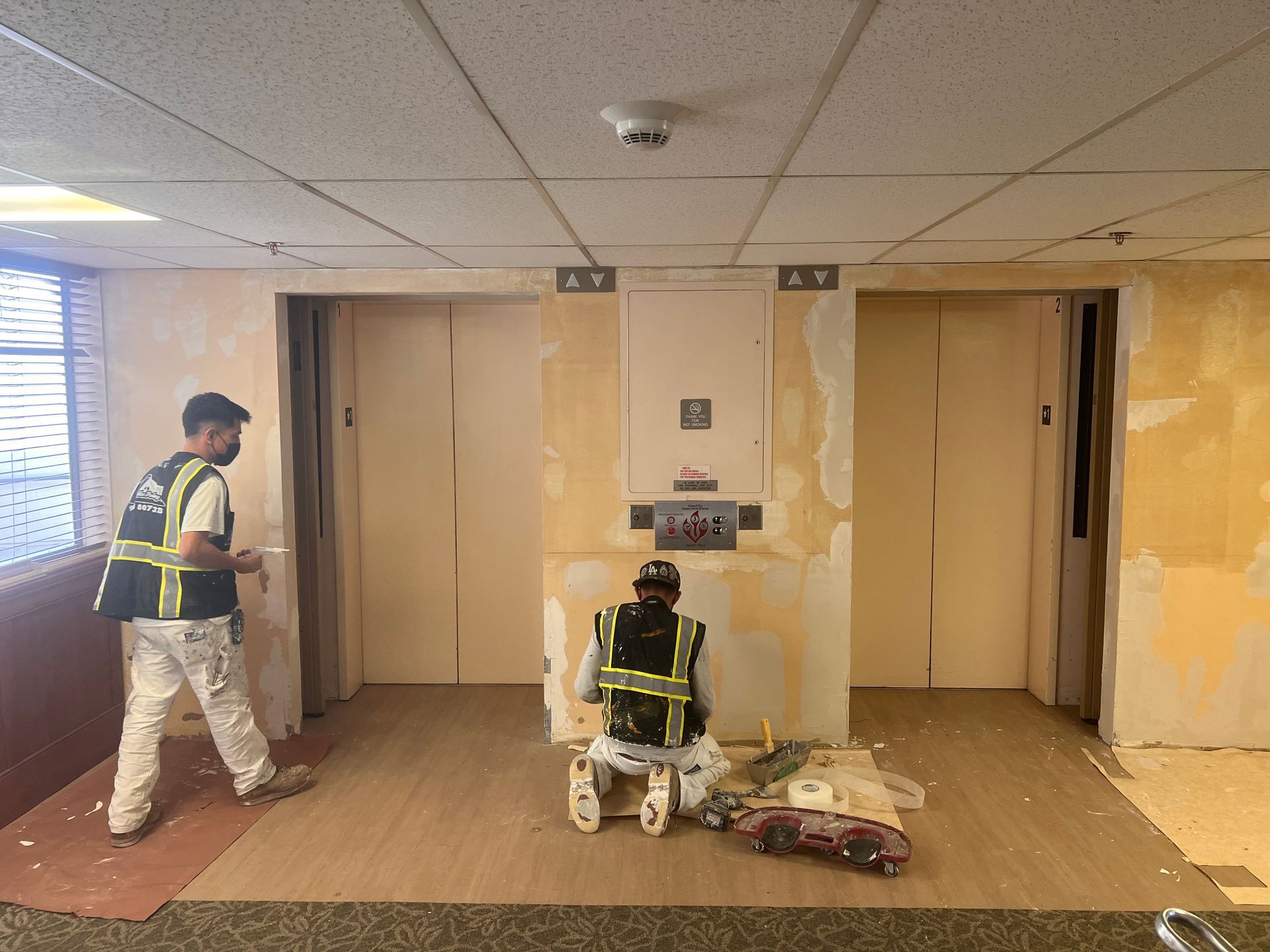 Two workers in high-visibility vests renovate a lobby area with two closed elevator doors and beige, unfinished walls.