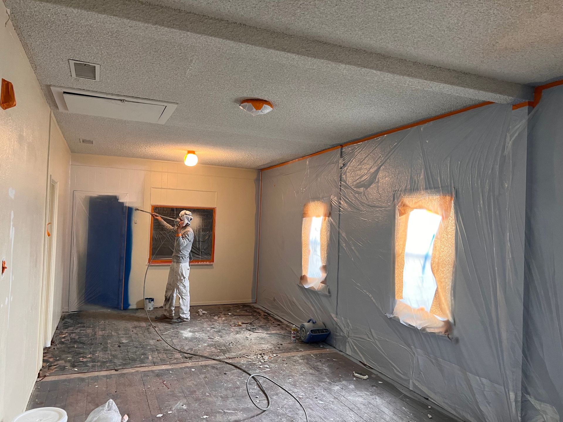 A person uses a paint roller on a wall in a room under renovation, with plastic sheeting covering walls and windows.