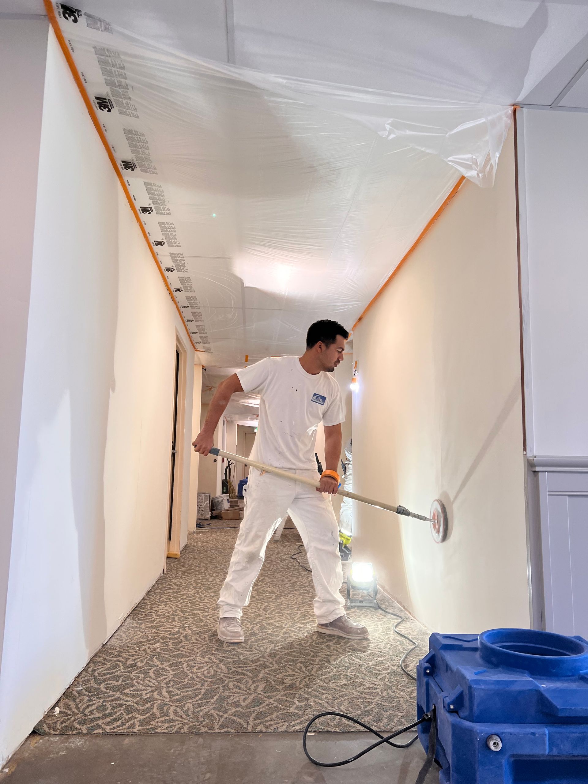 A person wearing white work clothes uses a long-handled sander to smooth a wall in a hallway covered in plastic sheeting.