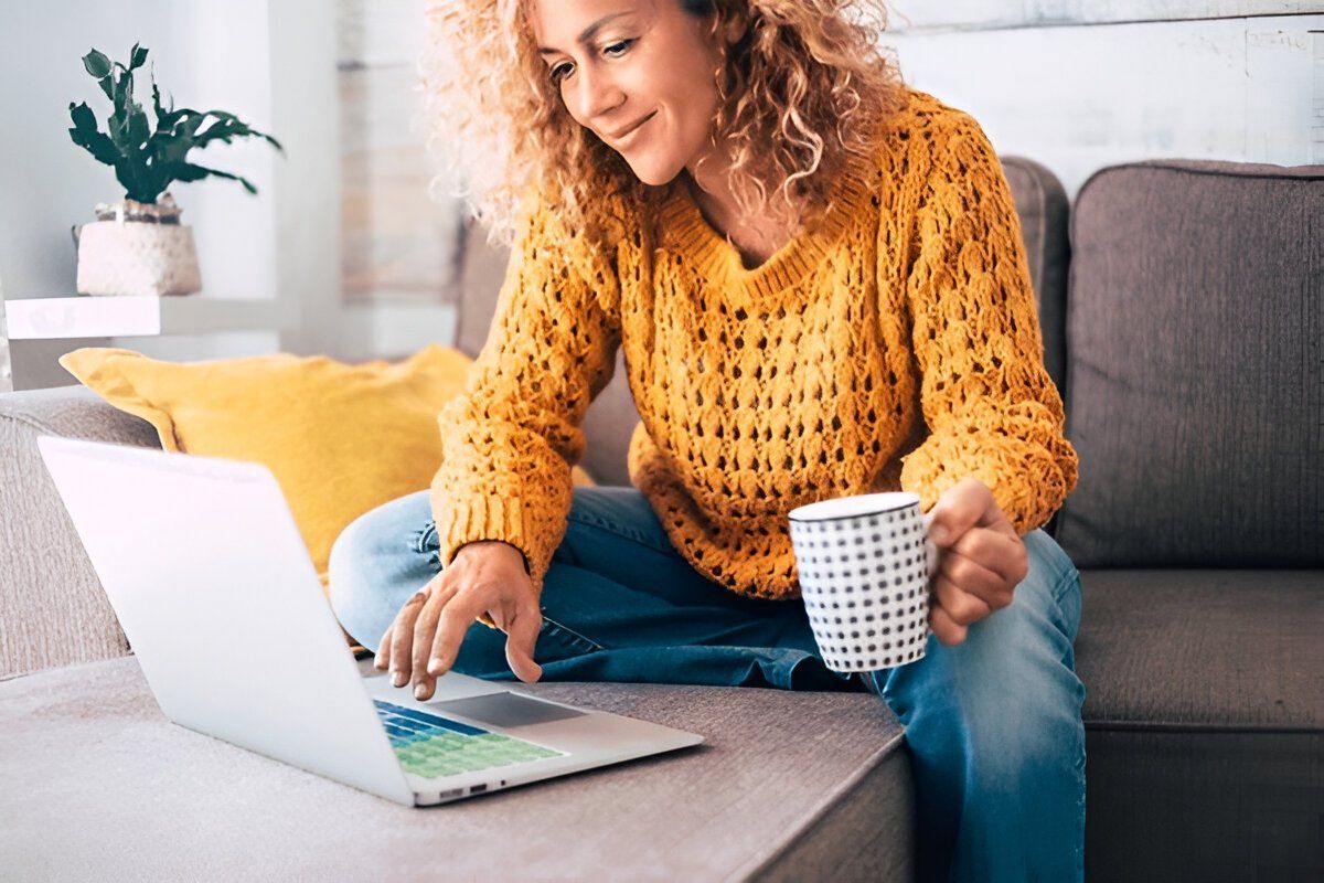 A woman is sitting on a couch using a laptop computer while holding a cup of coffee.