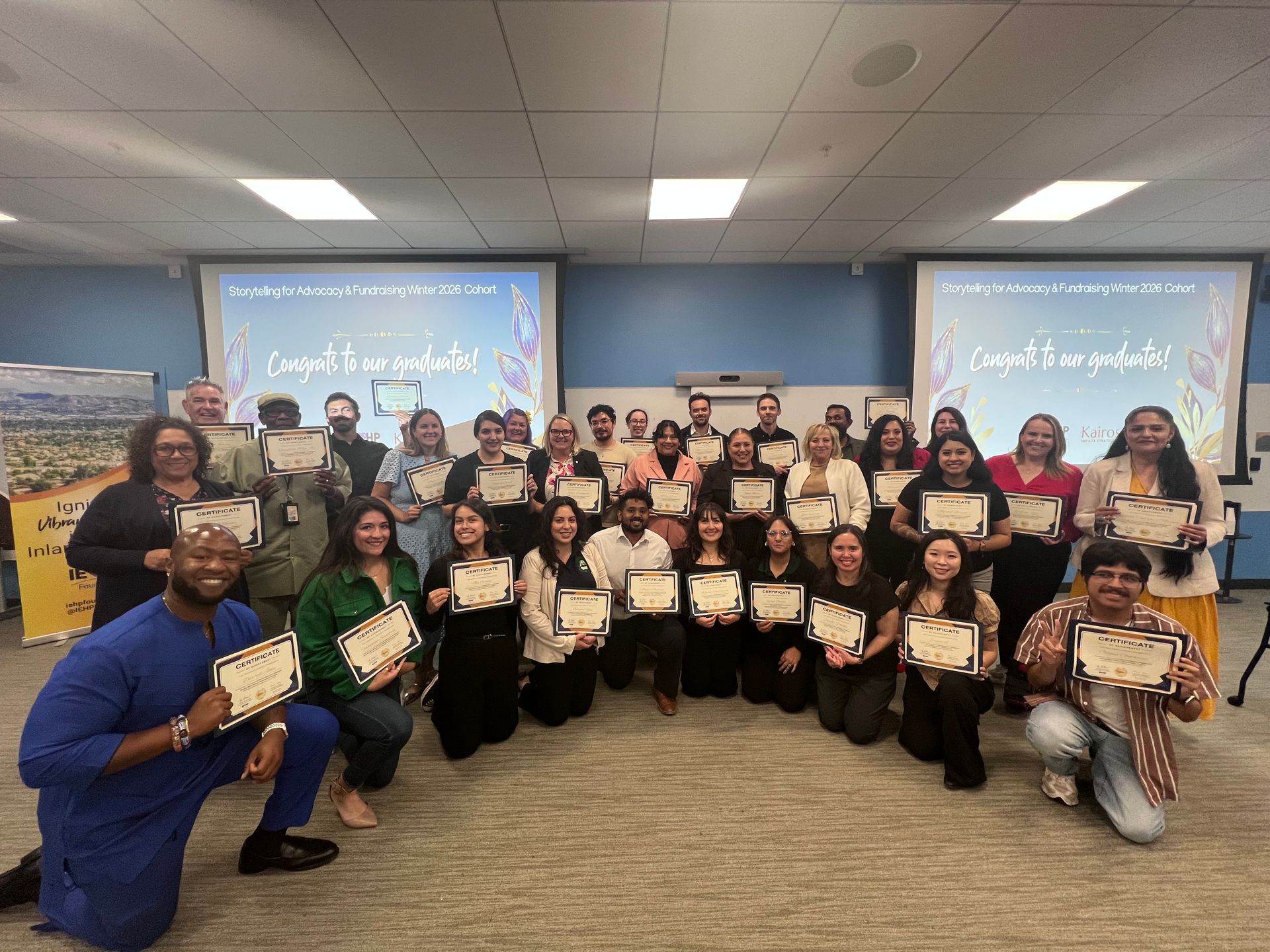 Group of people posing in a classroom, holding certificates and smiling in front of two projection screens